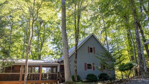 A cabin with a deck, surrounded by tall trees and a gravel path.