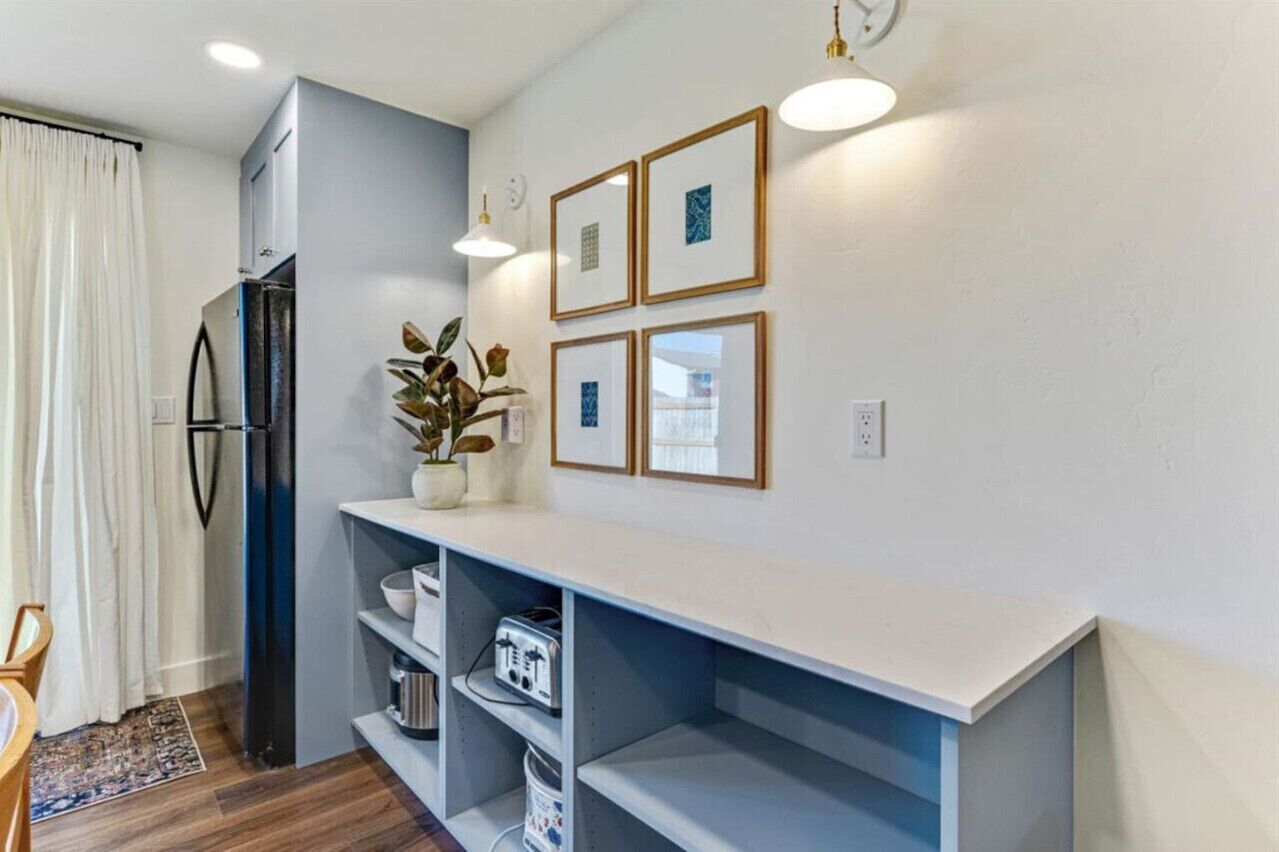Une cuisine moderne dotée d’un réfrigérateur encastré, d’un plan de travail blanc et d’une étagère. Des tableaux encadrés ornent le mur et une plante en pot est posée sur le plan de travail.