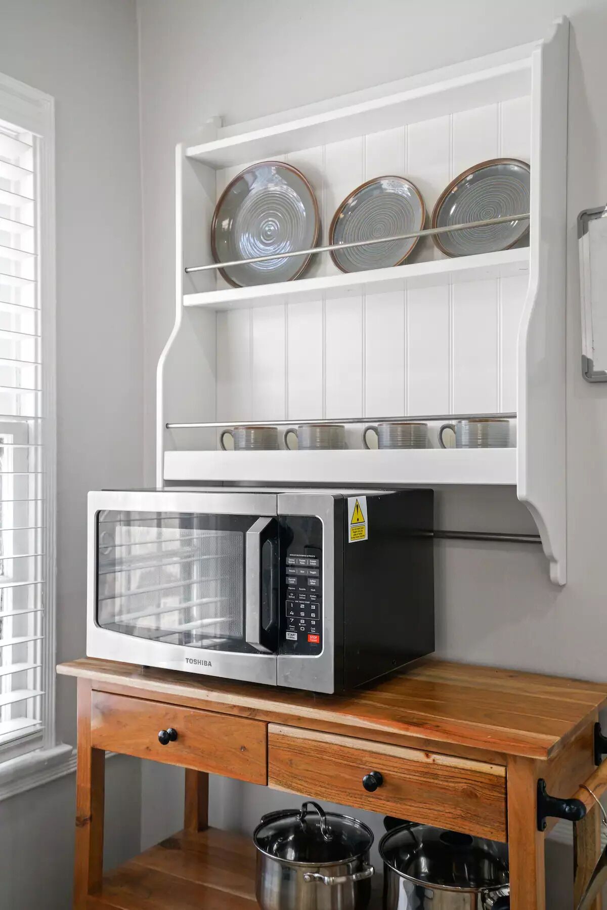 A kitchen with a microwave on a wooden counter, a shelf above holding plates and cups, and a window with blinds.