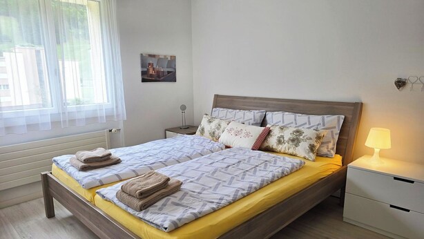 A double bed with a wooden frame, a bedside table with a lamp, and a window with sheer curtains.