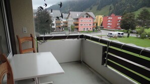 A balcony with a white table and chairs, overlooking a residential area with colorful buildings and a green landscape.