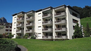A multi-story building with balconies, a paved driveway, and a grassy area with trees.