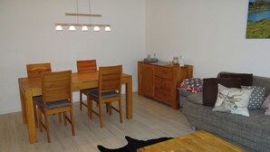 A living room with a wooden dining table and chairs, a sofa with patterned cushions, a wooden cabinet, and a painting on the wall.