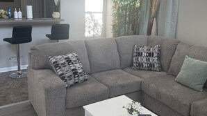 A living room with a grey sectional sofa, a white coffee table, and a patterned rug. There is a bar area in the background with a clock and decorative items. - Vacation Home in Delmar (Delmar)