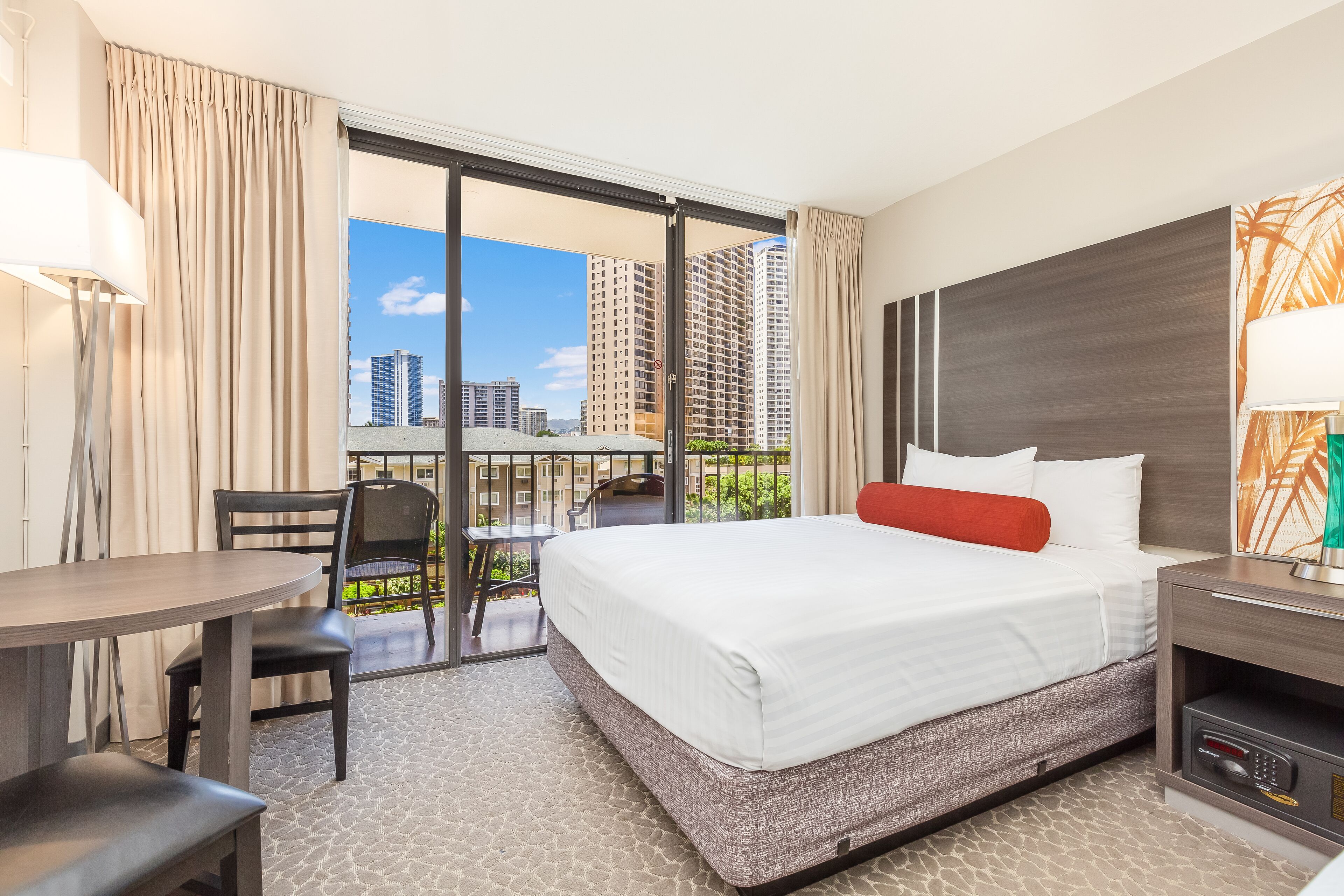 A hotel room with a large bed, a desk, and a chair. There is a balcony with a view of tall buildings.