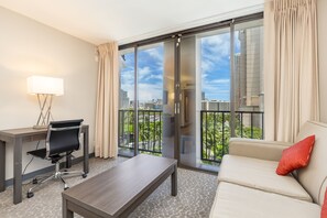 A modern hotel room with a large sliding glass door to a balcony, a desk with a chair, a sofa, and a lamp. - Castle at Palms at Waikīkī (Honolulu)