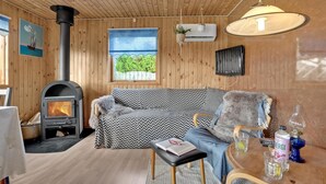 A cozy cabin interior with a wood-burning stove, a sofa, a small table with a book, and a window offering a view of greenery.