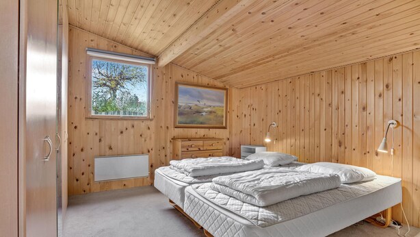 A room with wooden walls and ceiling, two beds with white bedding, a radiator, a painting, and a window with a view of trees.