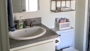 A bathroom with a sink, mirror, and a metal shelf above the toilet holding toiletries. - Peaceful European Cottage with (Ocala)