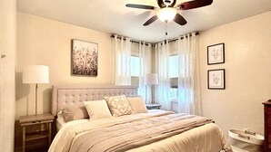 A bedroom with a bed, a ceiling fan, a nightstand, a lamp, and a window with curtains. - Charming bungalow in the heart of Southwest Colorado (Cortez)