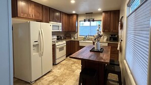 A compact kitchen with wooden cabinets, a white refrigerator, a microwave, and a stove. There is a window with blinds, a sink, and a wooden dining table with chairs. - Charming bungalow in the heart of Southwest Colorado (Cortez)