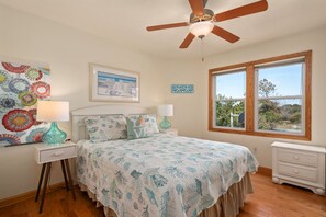 A bedroom with a bed, two bedside tables, a ceiling fan, a window with a view, and a wall with decorative items. - 123 Beach Therapy-Oceanside, private pool, hot tub, dog friendly, close to beach (Duck)