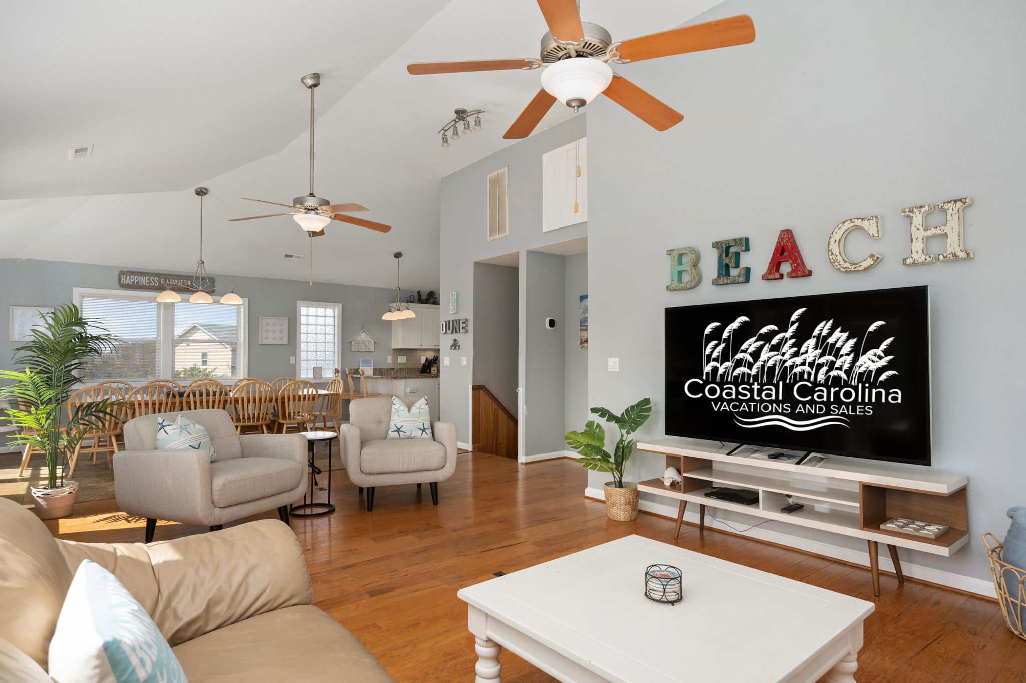 A spacious living room with a large flat-screen TV, wooden ceiling fans, and a cozy seating area with a sofa and armchairs.