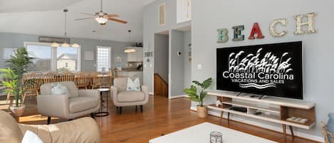 A spacious living room with a large flat-screen TV, wooden ceiling fans, and a cozy seating area with a sofa and armchairs.