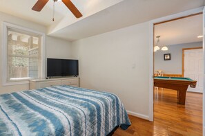 A bedroom with a bed, a television, a window with blinds, a ceiling fan, and a pool table in the adjacent room.