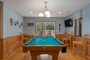A pool table with a green felt surface, surrounded by wooden chairs and a pool cue rack.