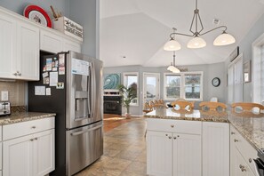 A modern kitchen with white cabinets, a stainless steel refrigerator, granite countertops, and a dining area with wooden chairs. - 123 Beach Therapy-Oceanside, private pool, hot tub, dog friendly, close to beach (Duck)