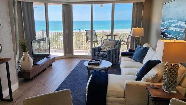 Ein geräumiges Wohnzimmer mit einem großen Fenster mit Blick auf einen Strand, ausgestattet mit einer Couch, Sesseln und einem Couchtisch.
