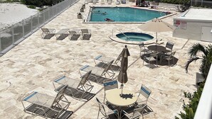 Ein Poolbereich mit Liegestühlen und Sonnenschirmen, im Hintergrund ein Strand und ein Gebäude mit Balkonen.