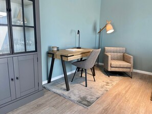 Quarto com escrivaninha de madeira, cadeira cinzenta, luminária e aparador com portas de vidro.