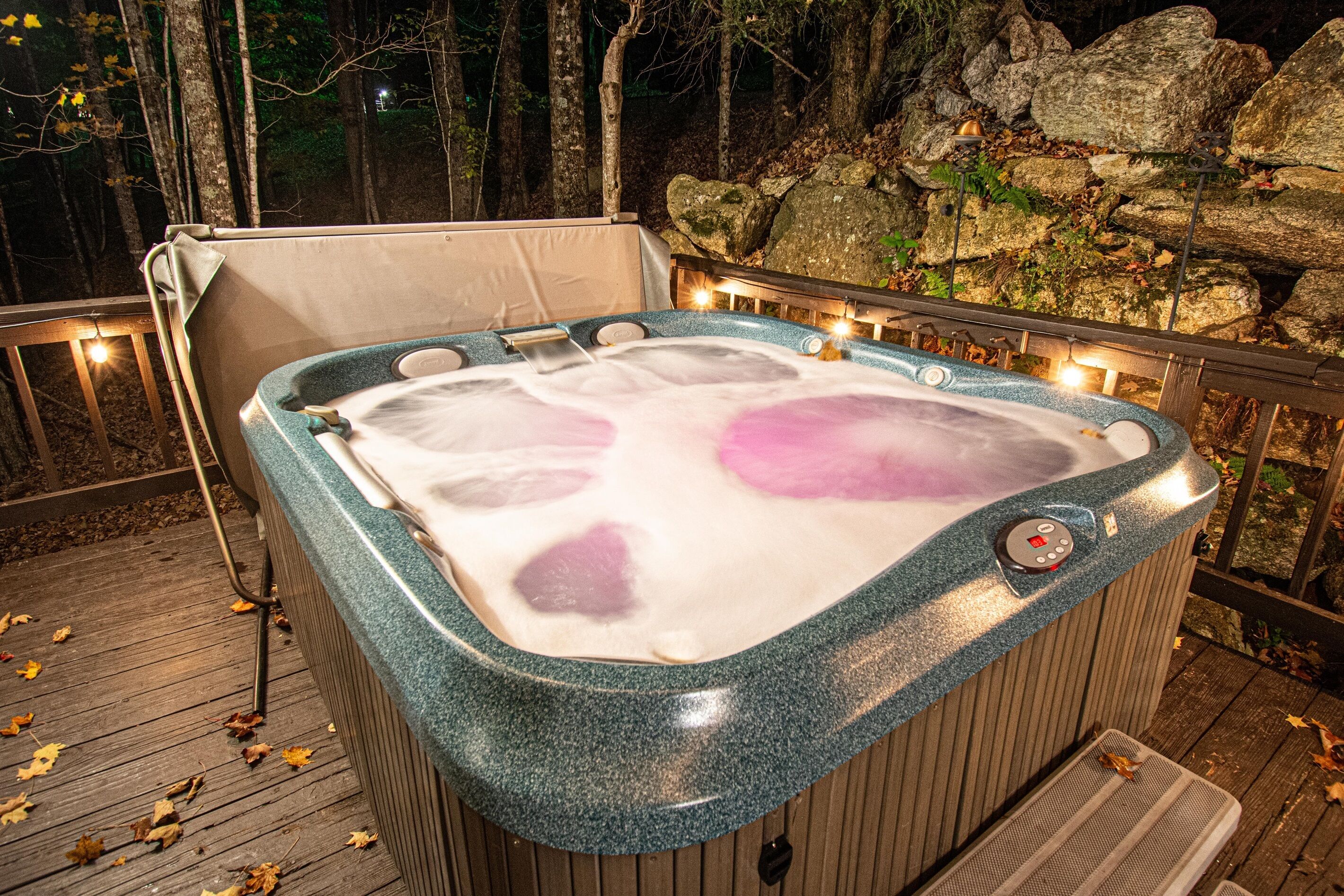 A hot tub with a control panel, surrounded by a wooden deck and a rocky wall.