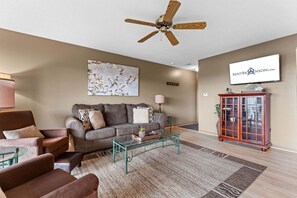A living room with a sofa, armchairs, a glass coffee table, a ceiling fan, a TV, and a cabinet.