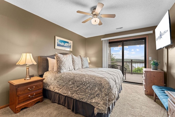 A bedroom with a bed, a nightstand, a lamp, a ceiling fan, a TV, and a balcony with a view.