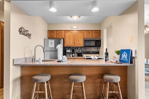 A kitchen with wooden cabinets, a granite countertop, a refrigerator, a microwave, a coffee maker, and a bar stool.