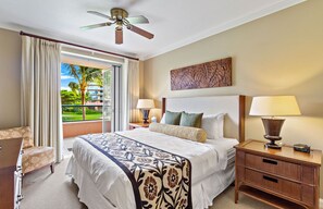 A hotel room with a large bed, two bedside tables with lamps, a ceiling fan, a view of a palm tree and building through a window, and a chair. - Maui Resort Rentals: Honua Kai – 2 Total Bedrooms + Den w/ Lush West Maui Mountain Views! (Lahaina)
