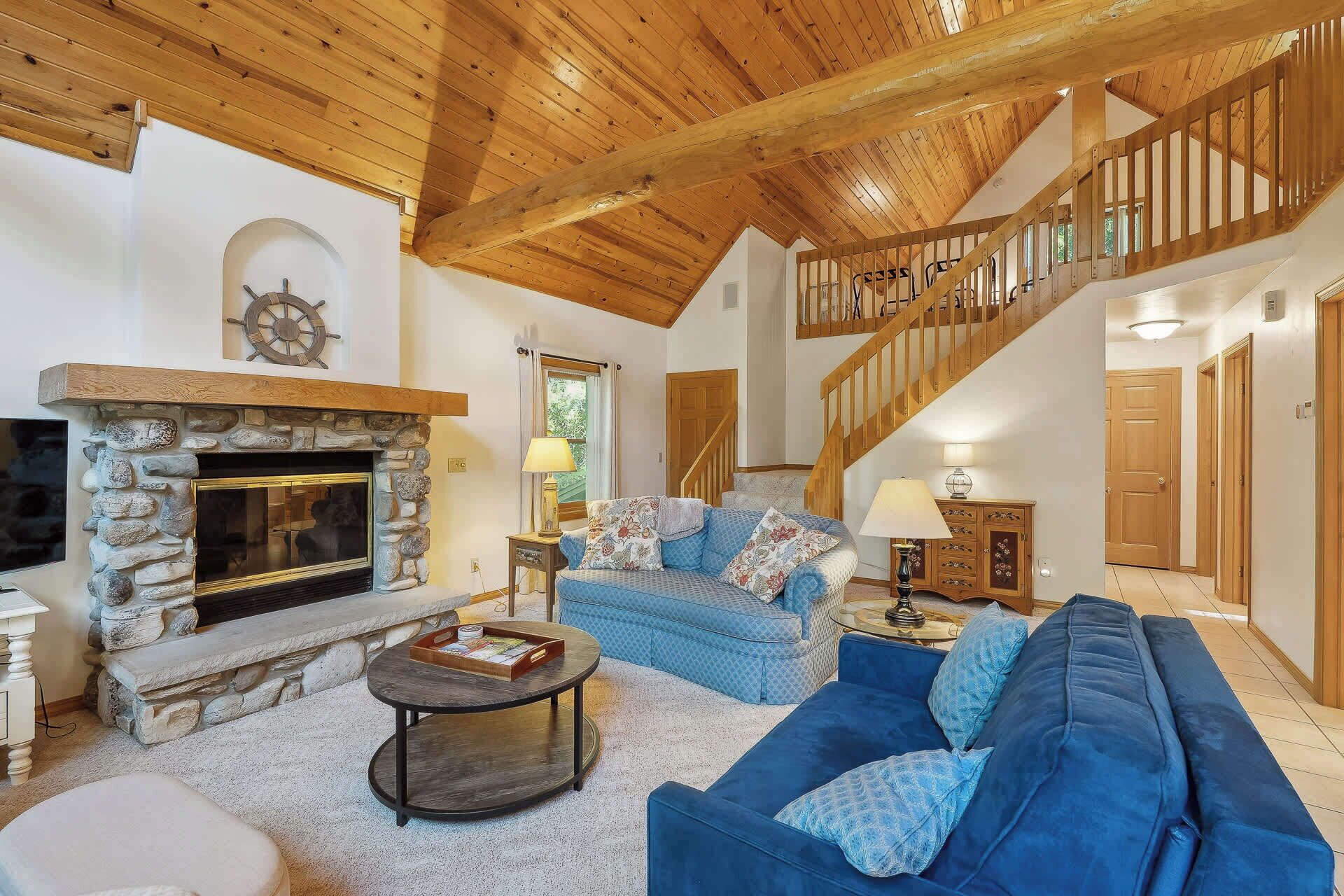 A spacious living room with a stone fireplace, a blue sofa set, a wooden staircase, and a view of the outdoors.