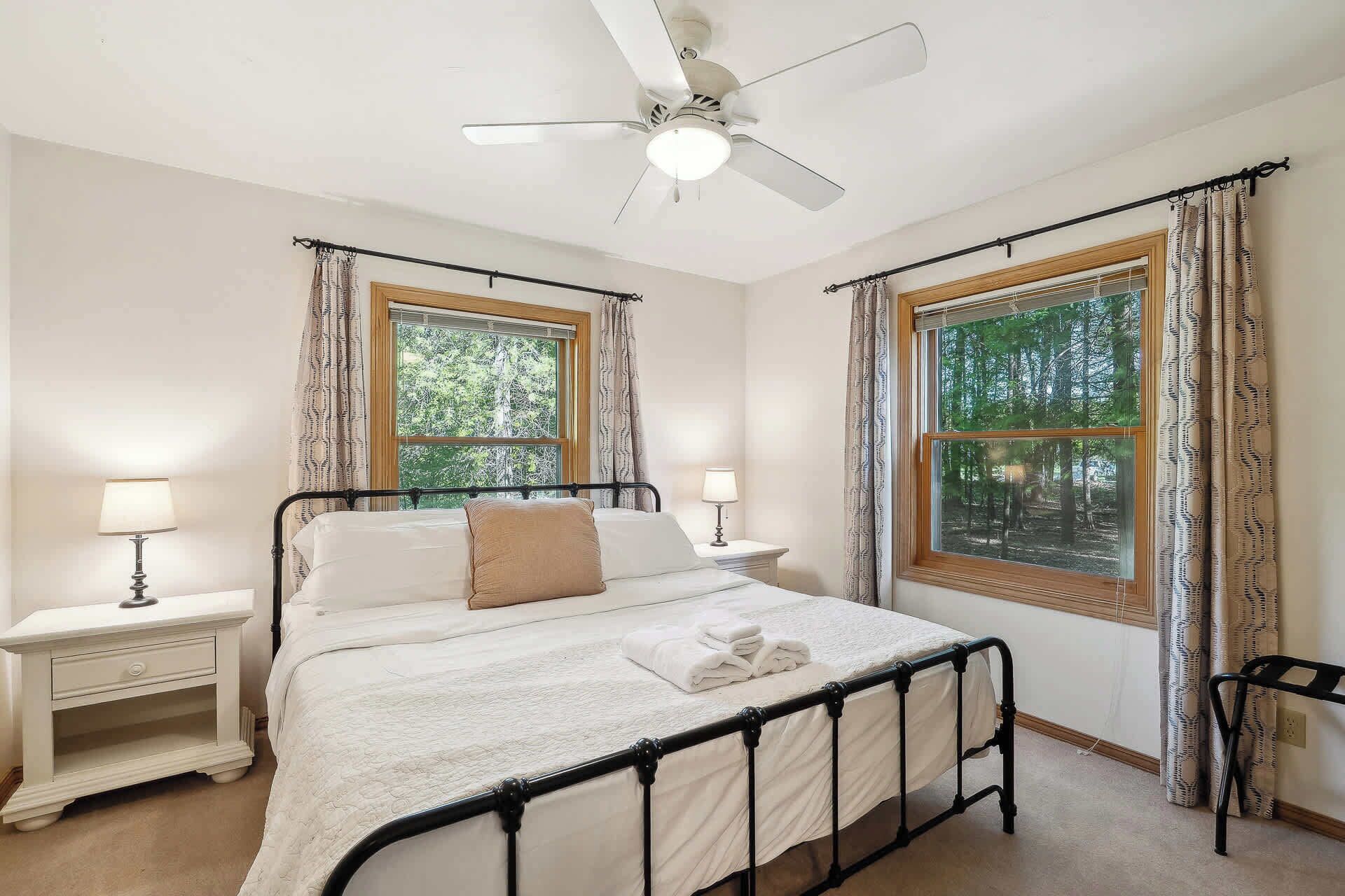 A bedroom with a bed, two bedside tables, a ceiling fan, and two windows with curtains.