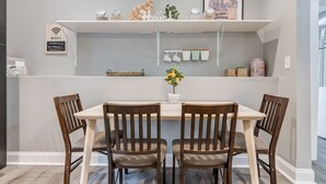 Un coin repas avec une table blanche, quatre chaises en bois, une plante en pot et une étagère où sont posés des bocaux et un tableau encadré.