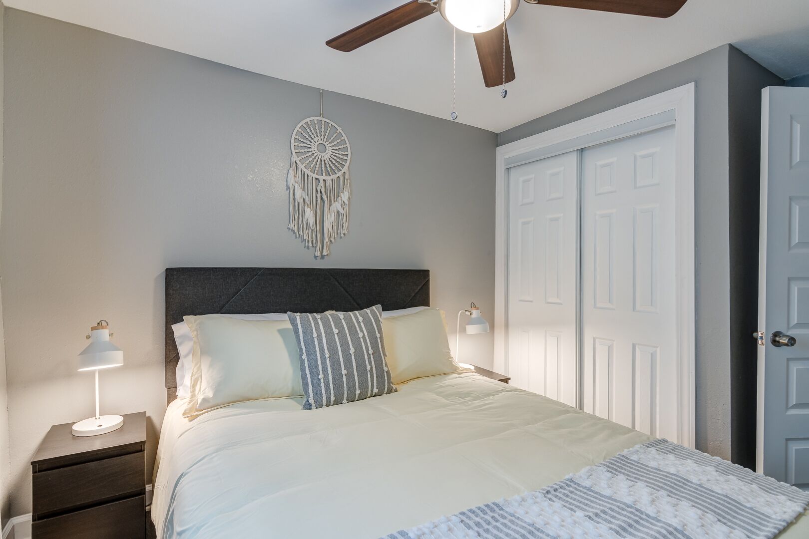 Une chambre à coucher avec un lit, un ventilateur de plafond, un attrape-rêves, une table de chevet avec une lampe et une porte.