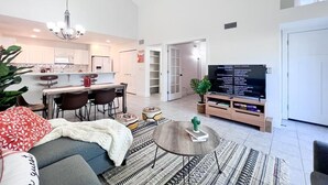 A modern living room with a sofa, coffee table, and a TV. There is a dining area with chairs and a kitchen in the background. - Southwestern Serene Oasis located in Chandler, featuring a community pool
 (Chandler)