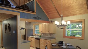 A spacious kitchen with wooden cabinets, a dining area with a table and chairs, and a large window with a view of trees. - Cozy Chalet in the Heart of Northern Michigan (Williamsburg)