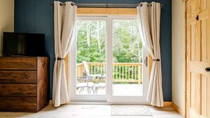 A room with a wooden dresser, a television, and a sliding glass door leading to an outdoor deck with a view of trees. - Cozy Chalet in the Heart of Northern Michigan (Williamsburg)