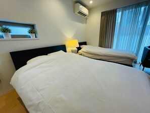 A hotel room with two beds, a bedside table with a lamp, an air conditioning unit, a window with curtains, and a small shelf with plants. - SALON Shibuya 5B (Tokyo)