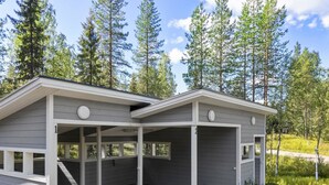 Eine kleine, zweistöckige Holzhütte mit einer Veranda und einer Garage, umgeben von hohen Bäumen.