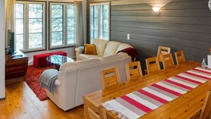 Ein gemütliches Wohnzimmer mit einer Couch, einem roten Hocker, einem Holzesstisch mit Stühlen und großen Fenstern mit Jalousien.