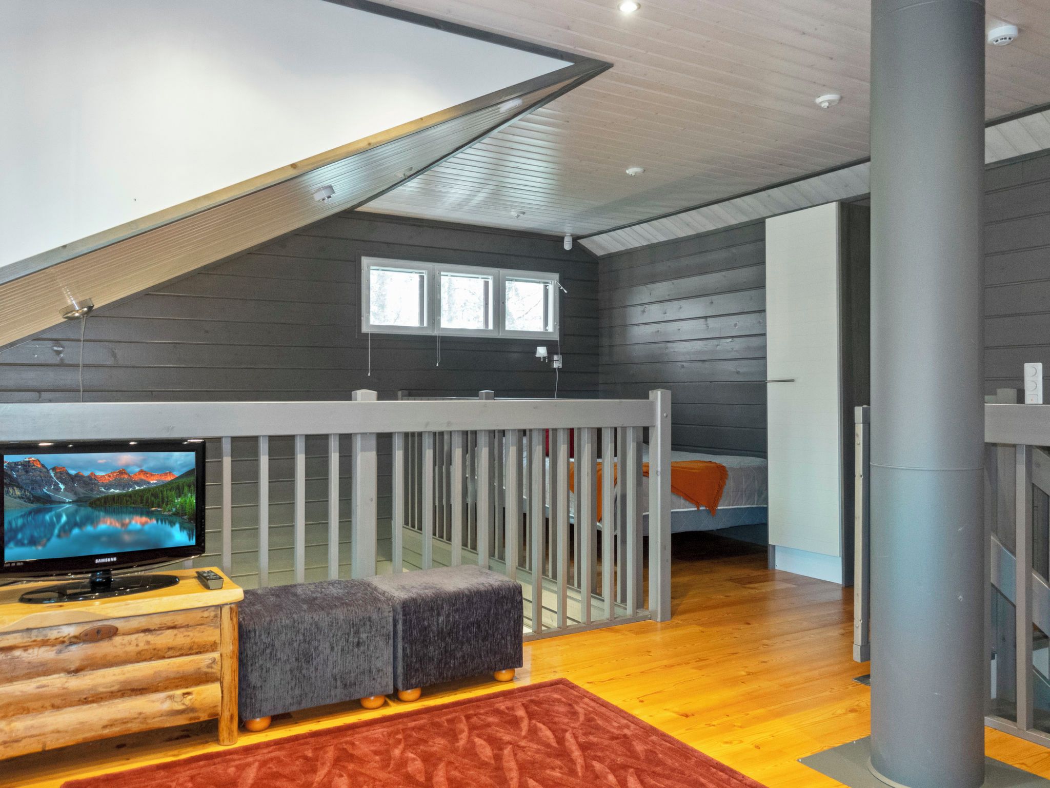 A modern interior with a wooden floor, a TV on a wooden stand, a grey bench, and a window with blinds.