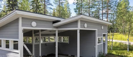 Eine zweistöckige Holzhütte mit überdachter Veranda und einem kleinen angebauten Garagengebäude, umgeben von einem Wald.