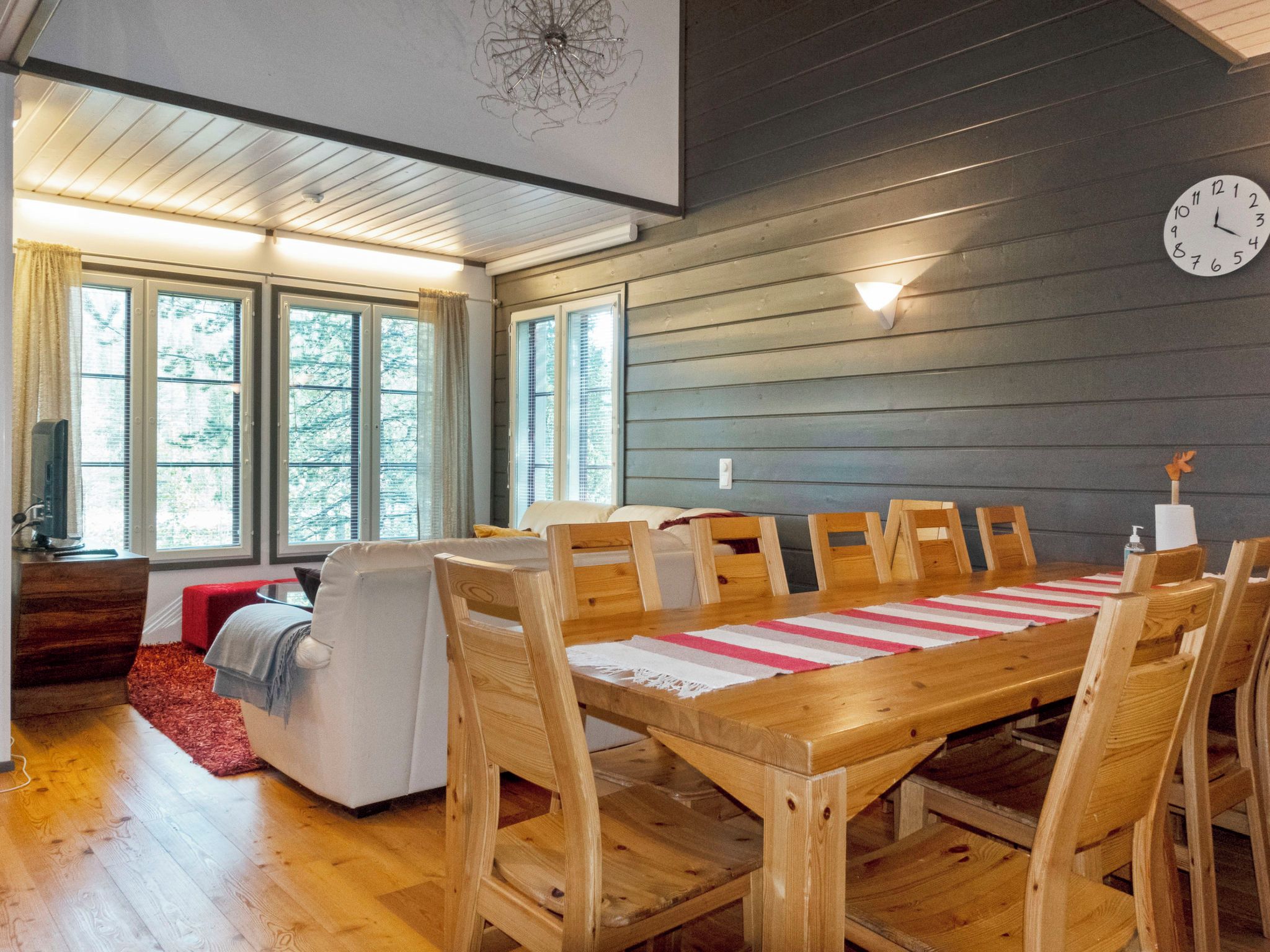A dining area with a wooden table and chairs, a clock on the wall, and large windows with blinds.