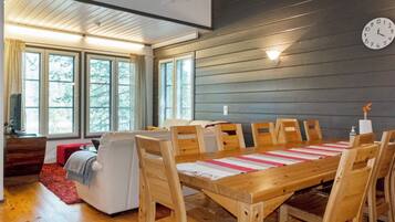 A dining area with a wooden table and chairs, a clock on the wall, and large windows with blinds.