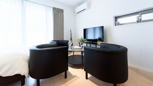 A modern living room with a bed, two black armchairs, a small round table, a TV, and a window with curtains.