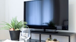 A modern living room with a flat-screen TV on a black stand, a glass table with a wine glass, and two potted plants.