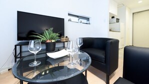 A modern living room with a glass table, two wine glasses, a potted plant, a flat-screen TV, and black leather sofas.