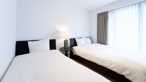A hotel room with two beds, a bedside table with a lamp, and a large window with curtains.