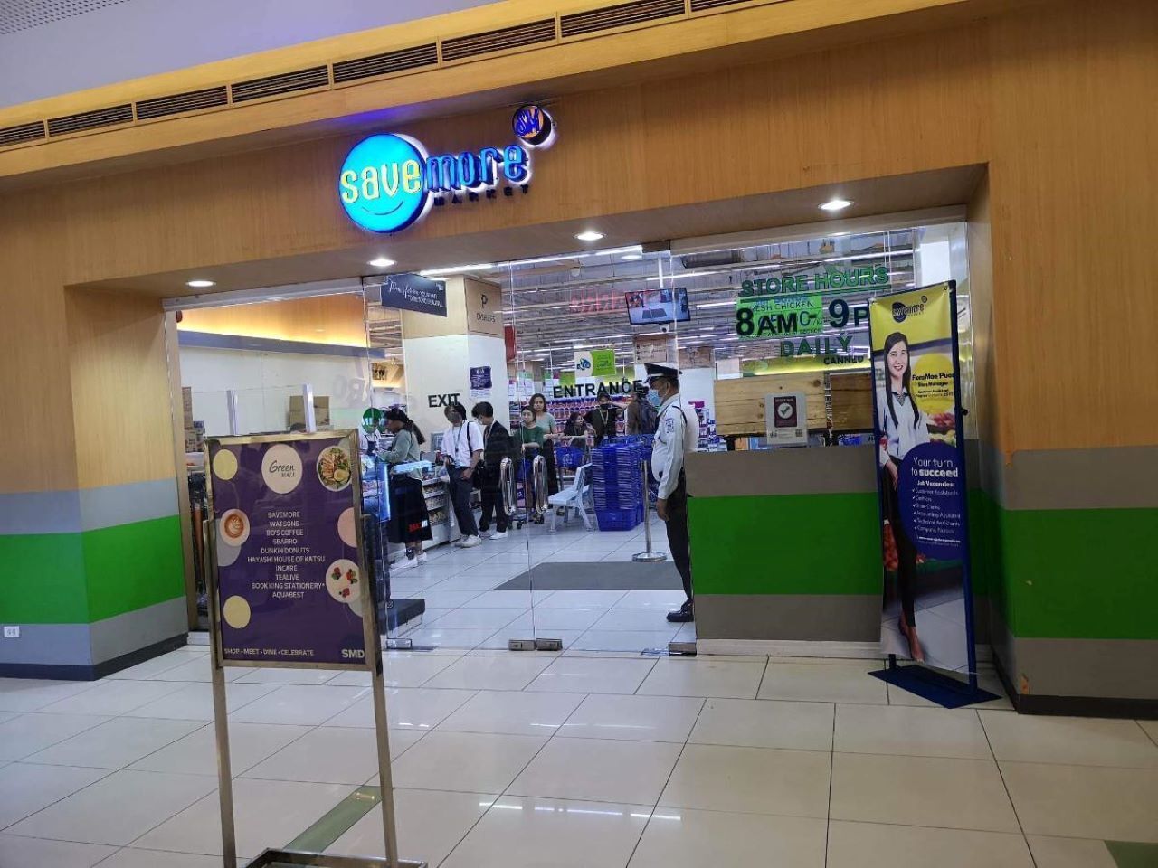 A food court area with a 'SAVEMORE' sign, a 'STORE HOURS' sign, and a 'Green Menu' sign.