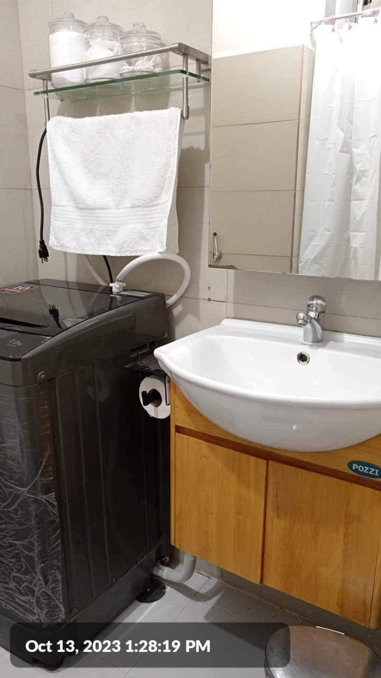 Une salle de bain avec une poubelle noire, un lavabo blanc et une étagère en verre au-dessus du lavabo.