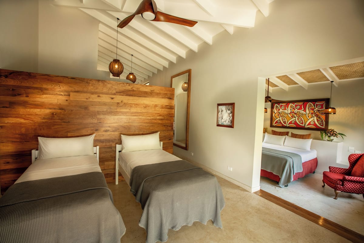 Quarto com duas camas, cabeceira de madeira, ventilador de teto, luminárias pendentes e uma poltrona vermelha.